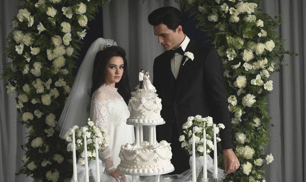 Priscilla Presley (Cailee Spaeny, in a white wedding dress and swept-back white veil), stands with Elvis Presley (Jacob Elordi, in a black tuxedo) stand behind their tiered white wedding cake under an arch of green leaves and white flowers, with Elvis looking downward and Priscilla looking directly into the camera, in Sofia Coppola’s Priscilla