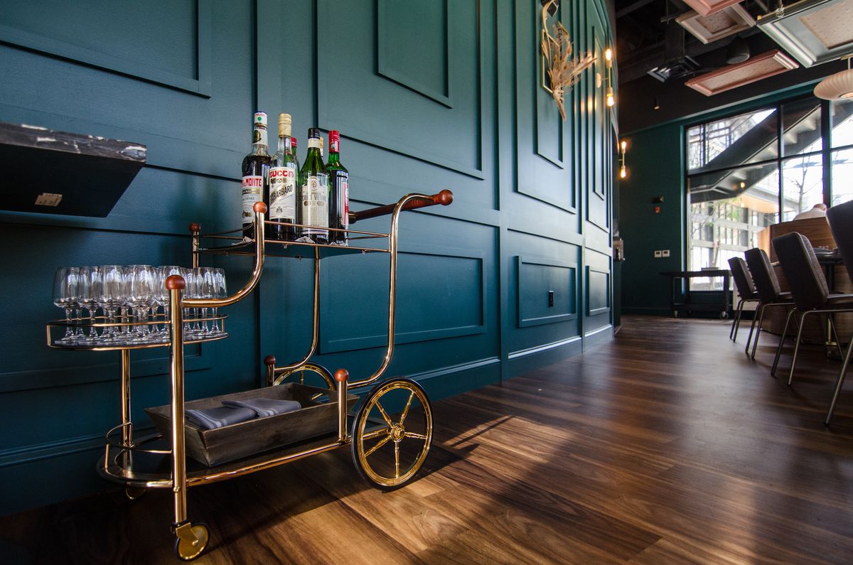 A small golden cart is parked along a dark teal wall of a restaurant, packed with bottles of amari and glasses.