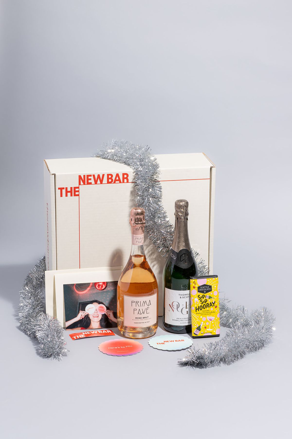 A gift set from the New Bar with two bottles of sparkling nonalcoholic wine and a yellow chocolate bar in front of a white box wrapped in tinsel.