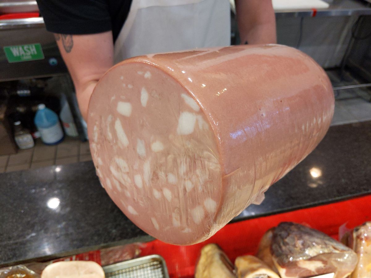 Two tattooed arms heft a large sausage over a cheese and meat case.