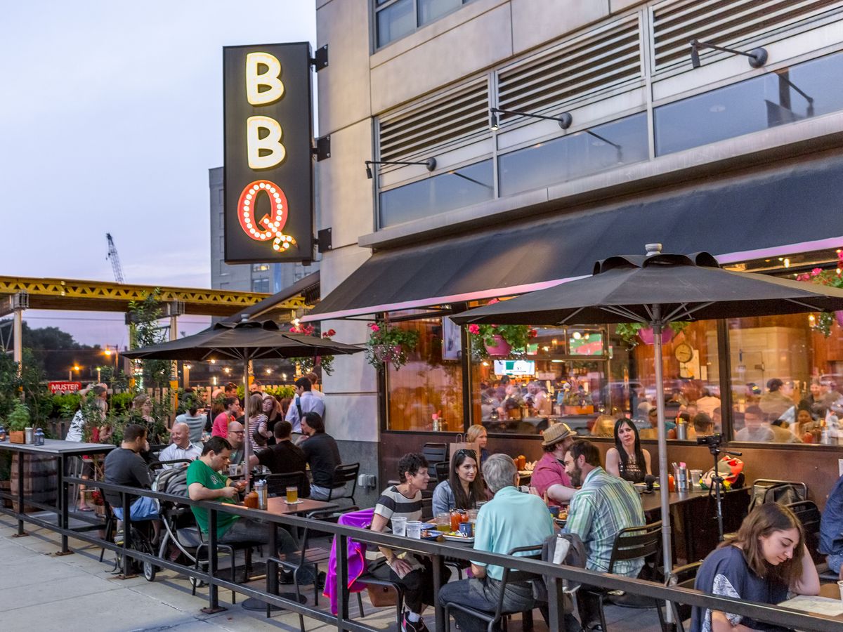 A sidewalk patio full of patrons outside of a restaurant with “BBQ” signage.