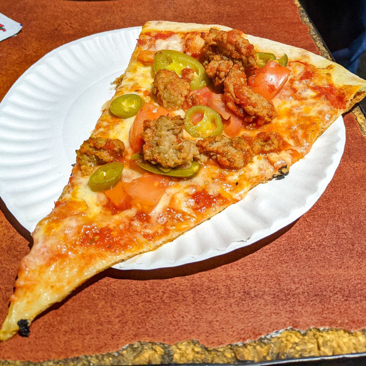 Slice of pizza with meatball, jalapenos and tomatoes with cheese and red sauce on a paper plate.