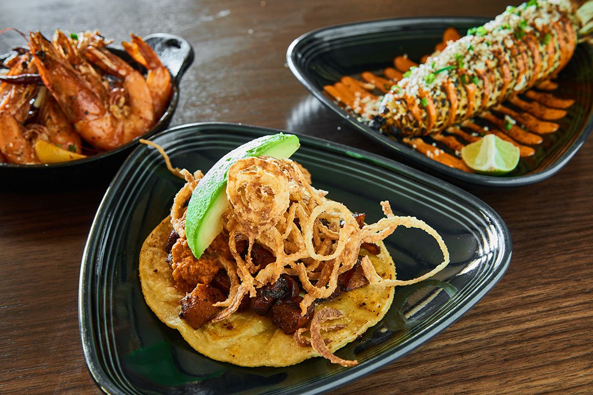 Three black plates rest on a dark wooden table. In the foreground, an avocado slice and crispy onion slices sit atop roasted butternut squash cubes on a corn tortilla. In the background on the right, a plate holds a lime wedge and a grilled corn on the cob drizzled with sauce, while the plate on the left holds shrimp.