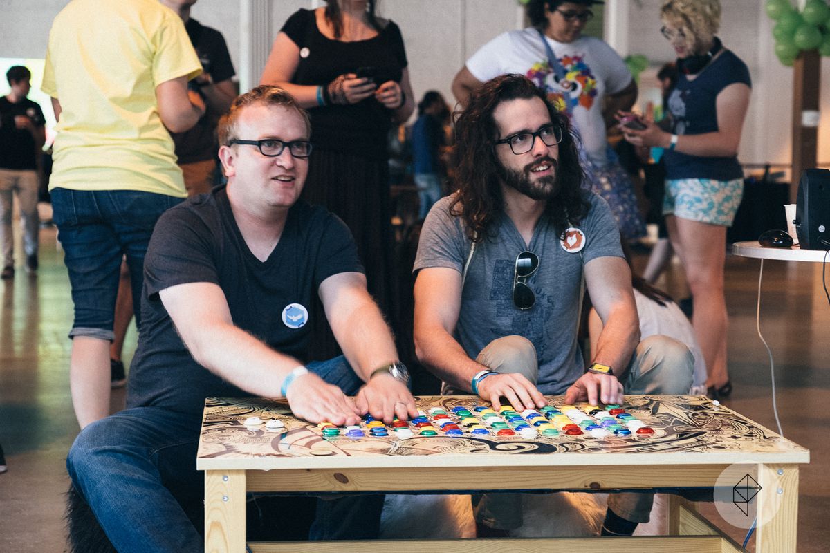 Two players sit in front of a hand-made wooden table with what can best be described as a ludicrous amount of buttons on it.