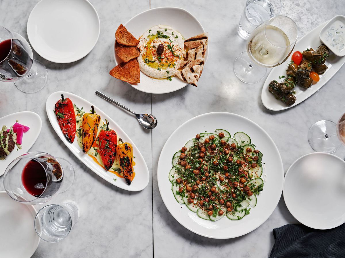 top-down view of an array of wine glasses and dishes on a white marble surface, including pitas and dips, roasted peppers, falafel balls, stuffed grape leaves, and a cucumber salad with crispy chickpeas