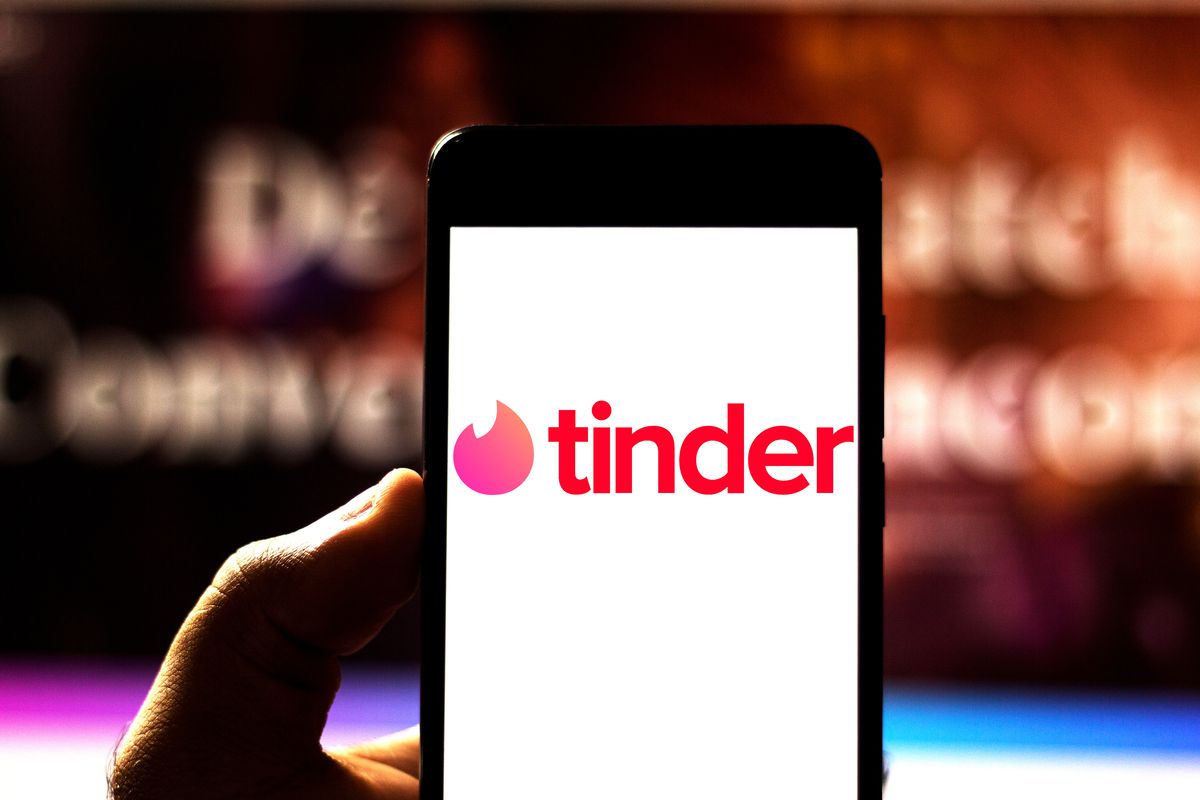 Photo illustration of a hand holding a mobile phone that is displaying the Tinder logo.