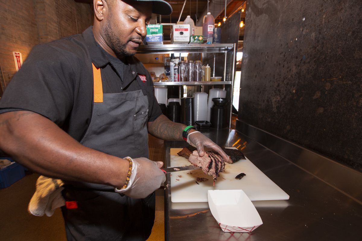 A male chef lifts sliced barbecue meat from a cutting board into a paper container.