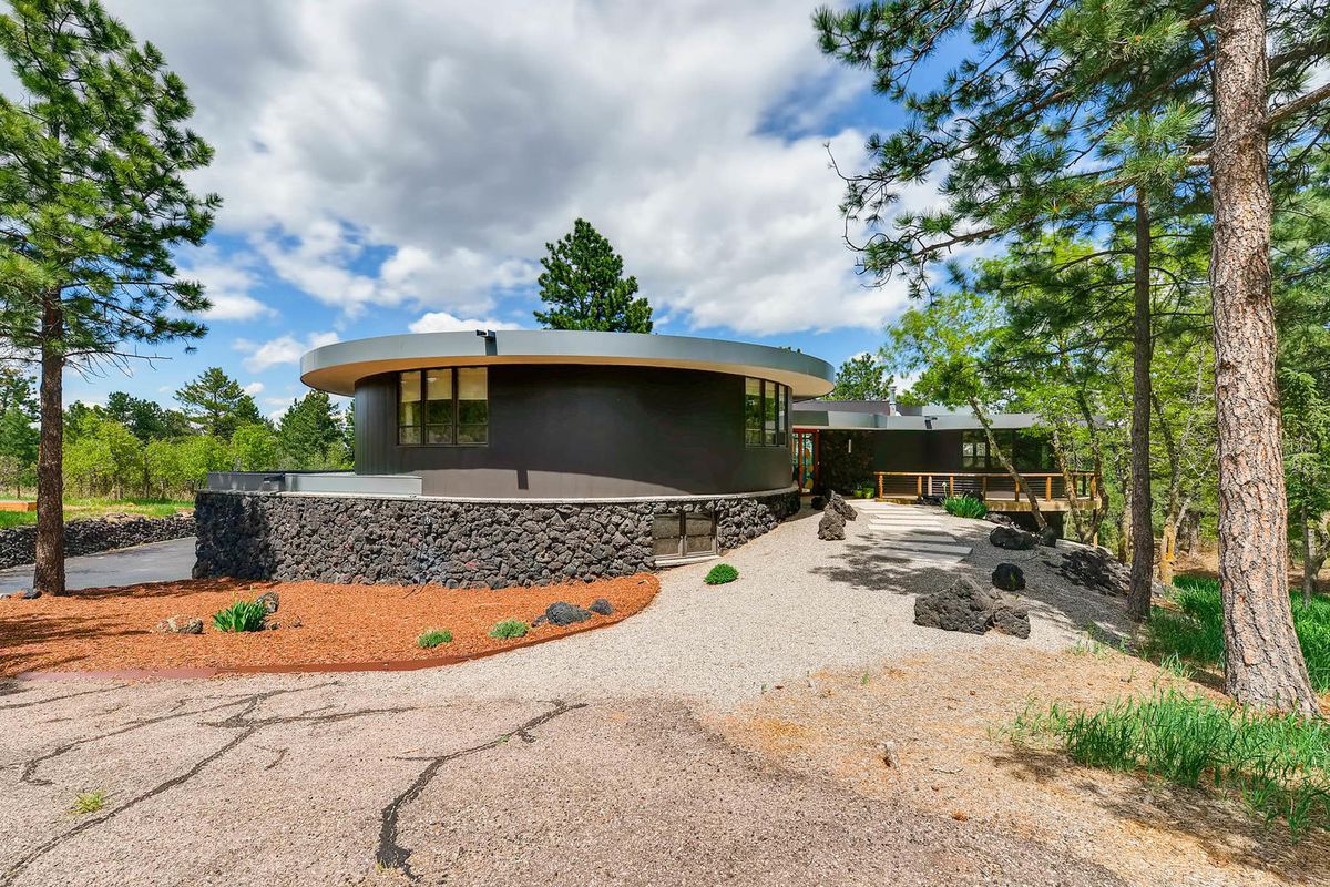 Exterior shot of home made up of large circular volume clad in wood and aluminum on the roof overhang. A stone wall surrounds a portion of it, and the home is set among trees. 