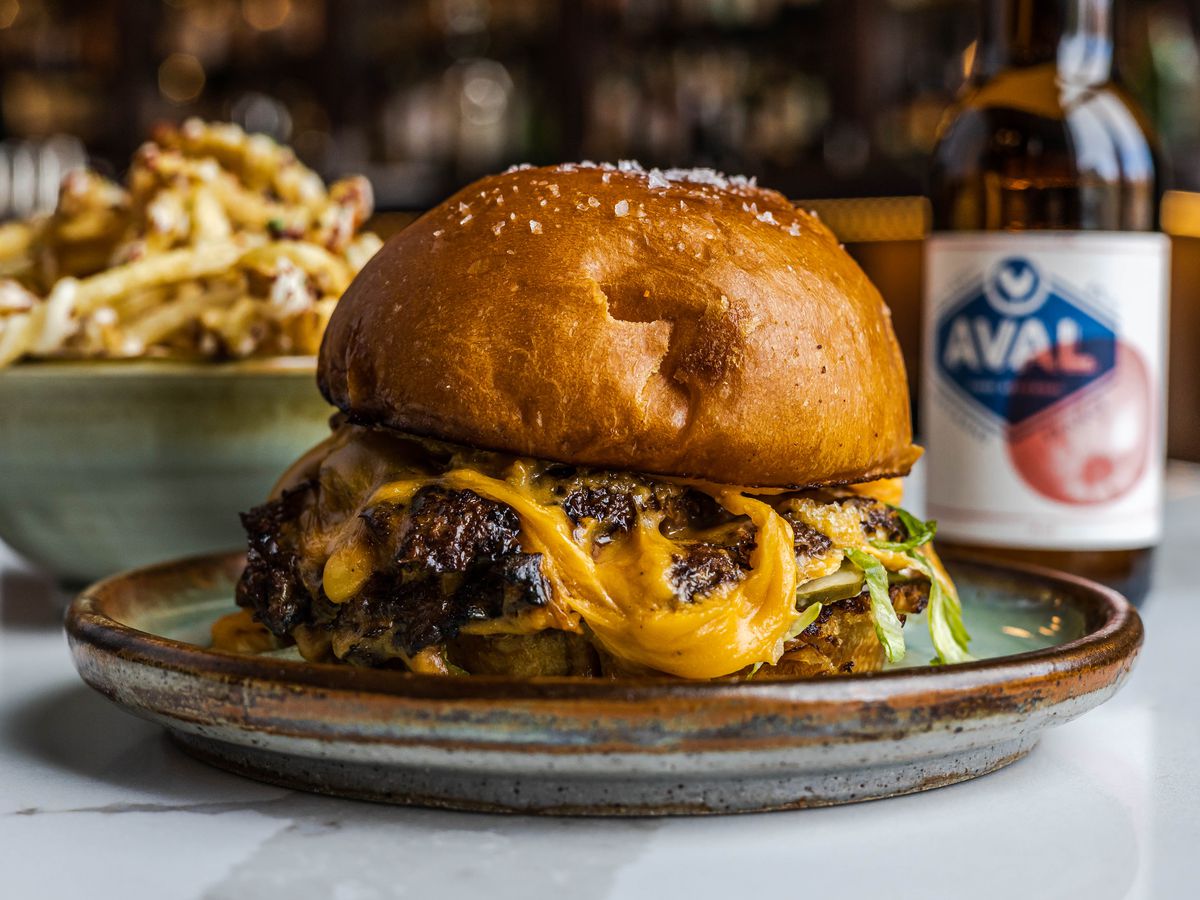 A cheeseburger in the foreground on a plate a bowl of fries to the left in the background and a brown bottle of beer to the right in the background from Bar Pigalle in Detroit, Michigan.