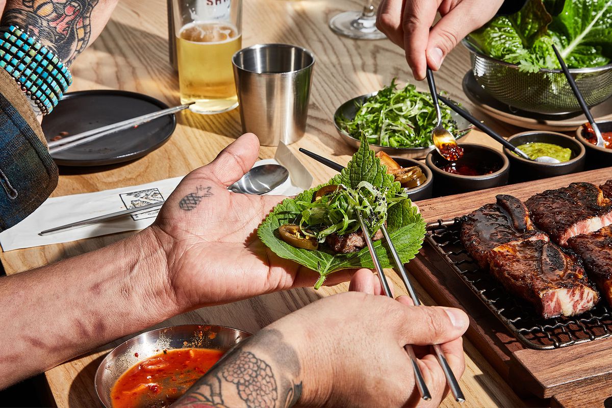 Two hands with tattoos use chopsticks to fill a large green leaf with bits of grilled meat and garnishes from a small tabletop grill. 