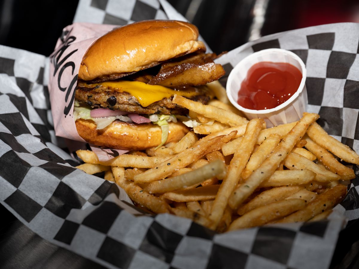 A vegan burger topped with vegan bacon with fries and ketchup in basket lined with white-and-black checkered paper.