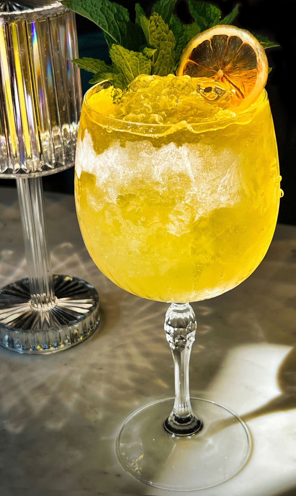 A wine glass filled to the brim with a yellow liquid and ice with a orange slice as a garnish.