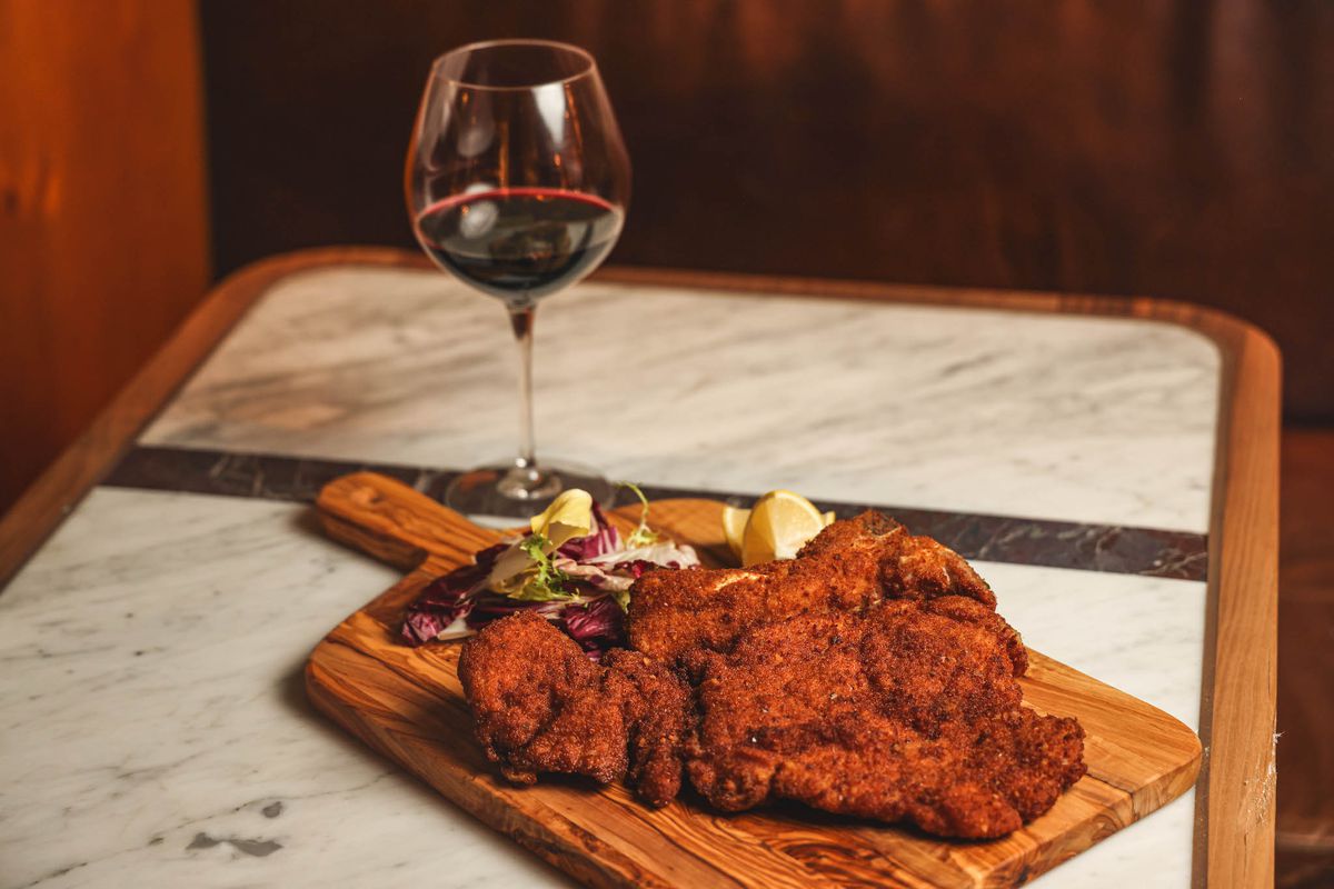 A cutting board with a breaded bone-in veal chop and glass of red wine next to it.