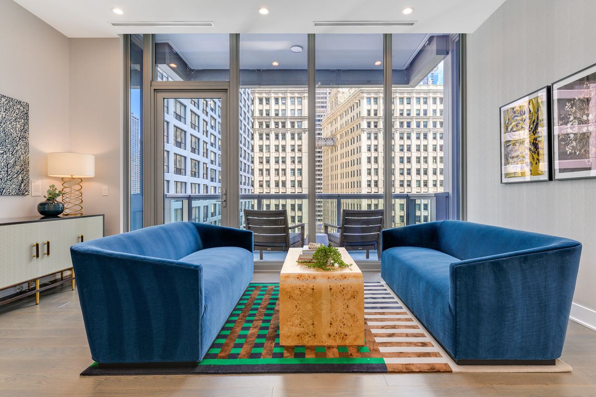 A living room with two blue couches on either side a wooden coffee table. The room connects to a balcony overlooking a cluster of taller buildings. 
