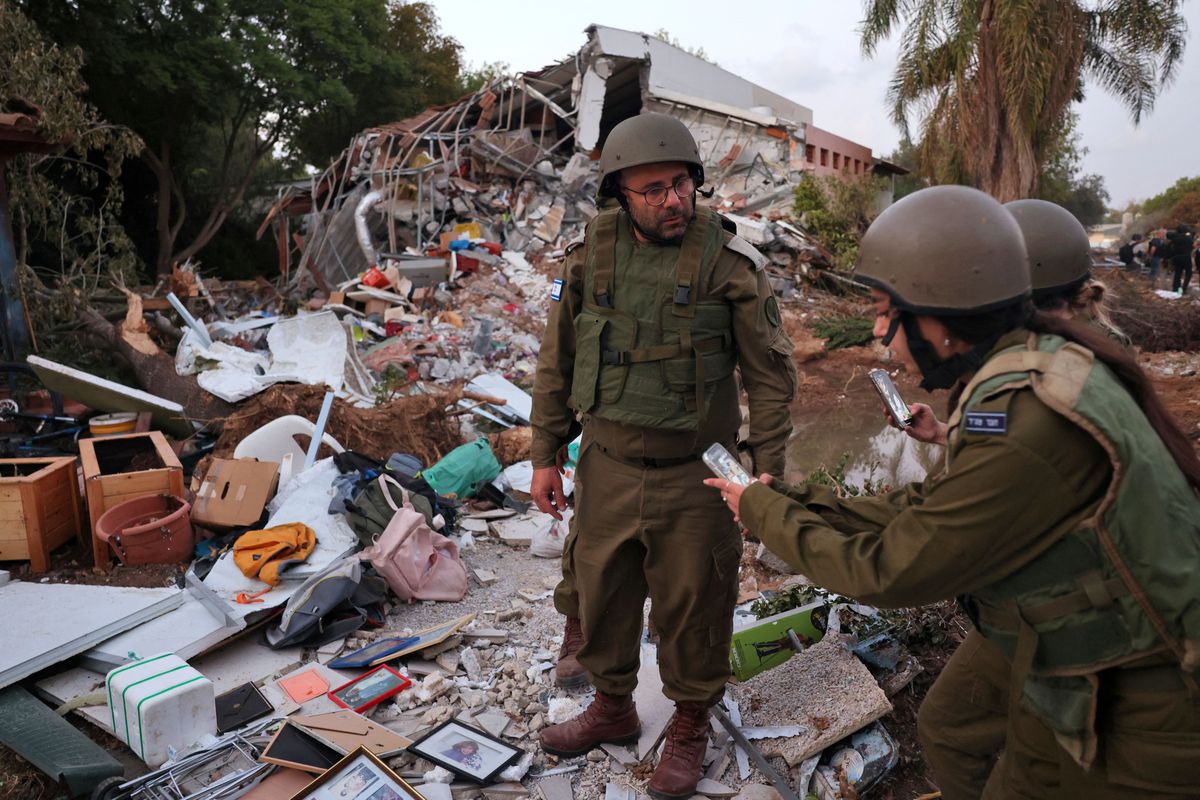 Three soldiers stand on a hillside with rubble of destroyed buildings and a torrent of household items covering the ground, taking photos.