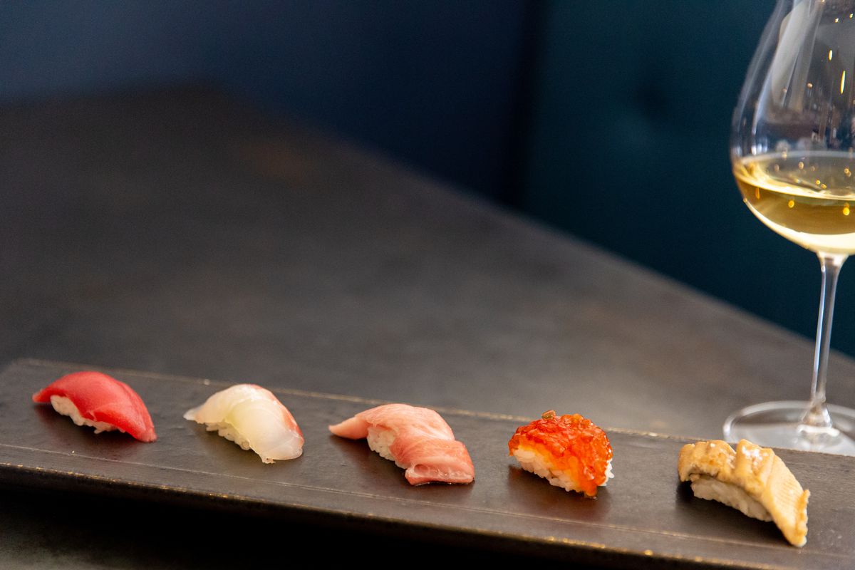 A slate board with five pieces of nigiri and a glass of white wine.
