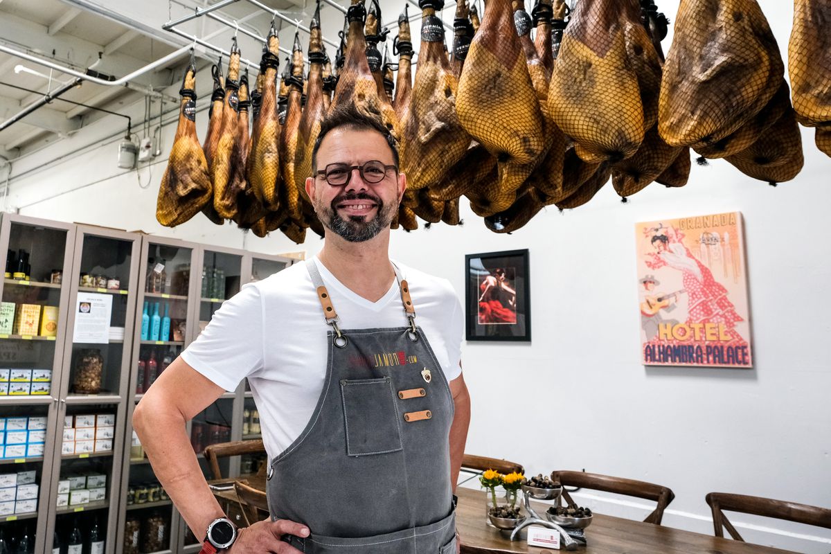 A chef stands in front of Iberico hams hanging from the ceilingg