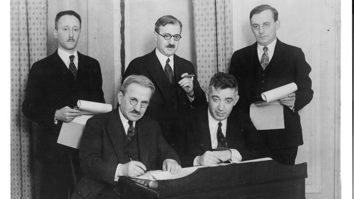 An old black and white photograph of five men, two sitting, with one signing a document.