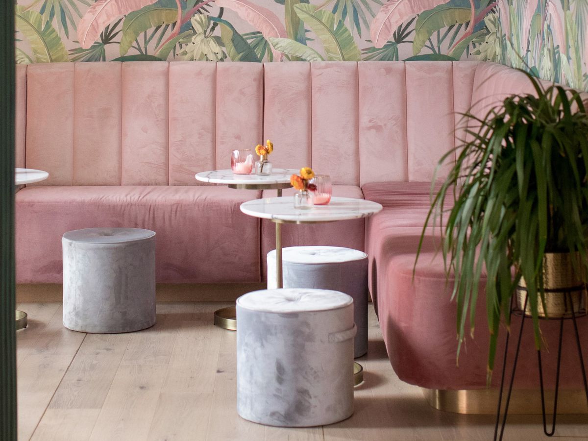 Pink banquettes in the corner of a bar room outfitted with tropical pink wallpaper. Short concrete-looking stools and small marble tables are scattered around, with a large plant in one corner.