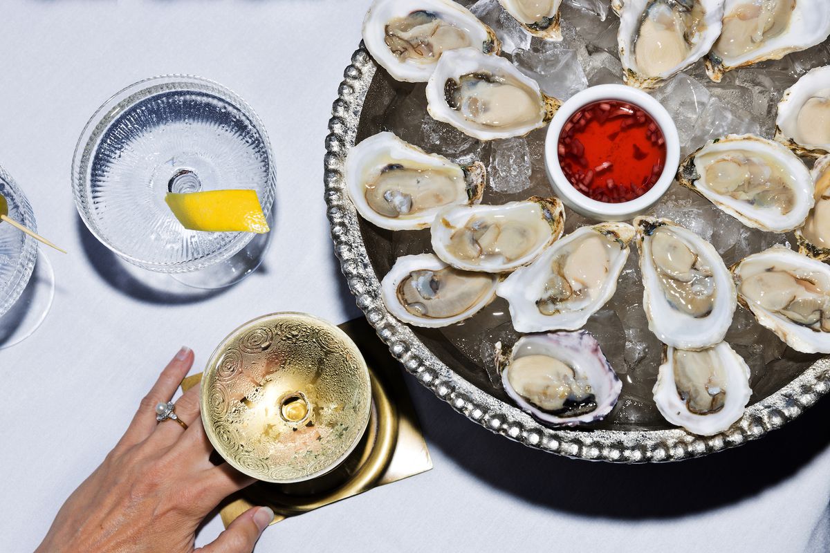Overhead flash shot of tray of raw oysters on ice with two drinks, one being held.