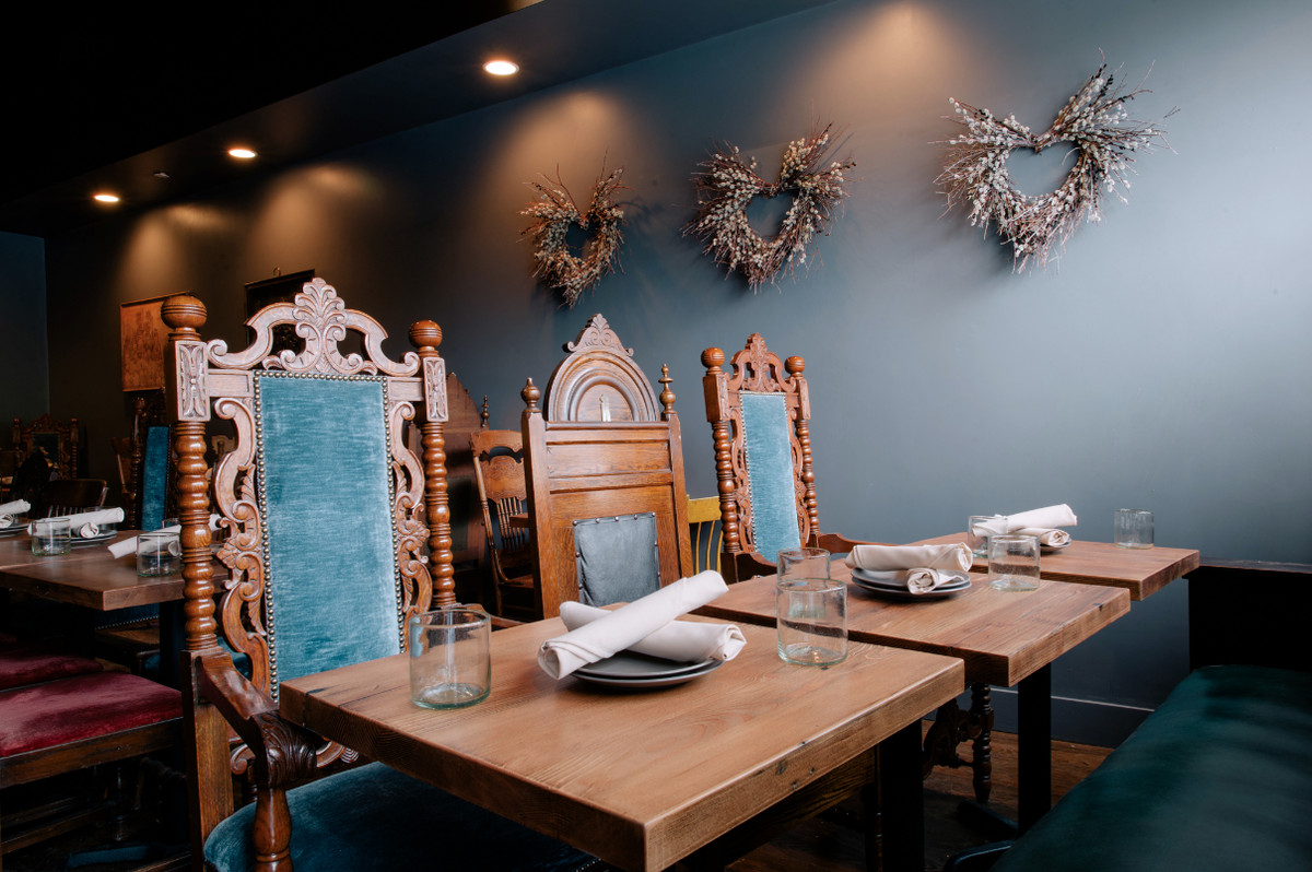 Finely carved wooden chairs with plush blue velvet padding line opposite wooden tables with a light blue wall in the rear at Bronwyn in Somerville.