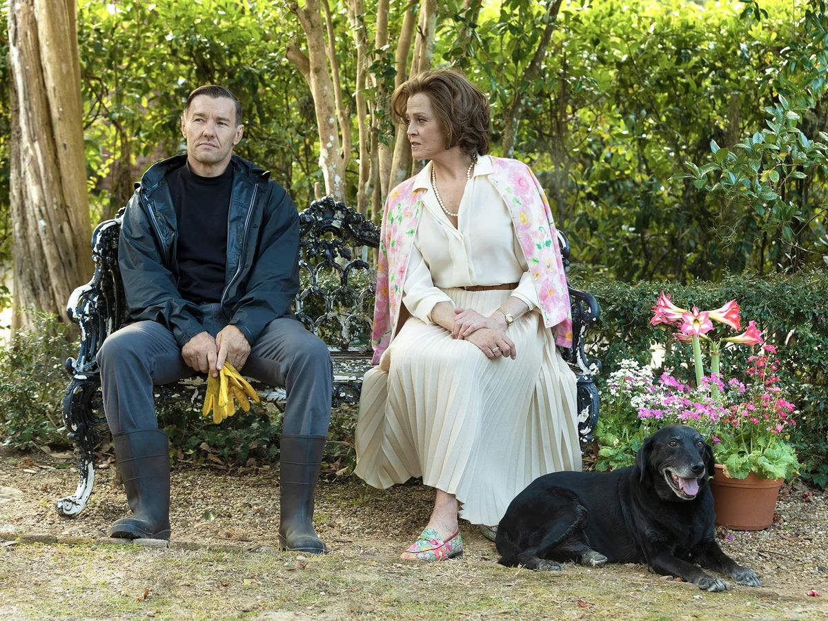 (L-R) Joel Edgerton&nbsp;and&nbsp;Sigourney Weaver&nbsp;sitting on a bench near an arrangement of potted flowers and a black dog in Master Gardener.