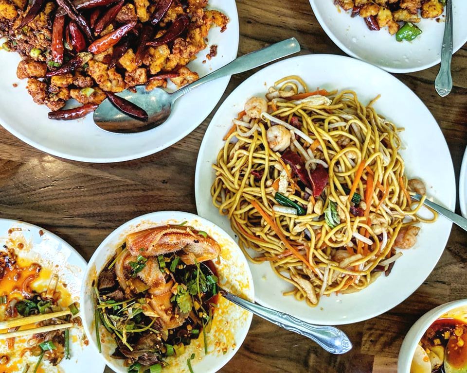 Overhead shot of a table of Chinese food, including lo mein, fried chicken, spicy wontons, and more
