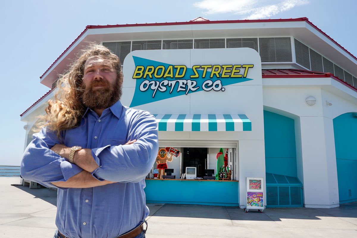 A man with long beard and wavy hair stands arms crossed in front of a white and teal seafood restaurant at daytime at Broad Street Oyster Co. in Huntington Beach.