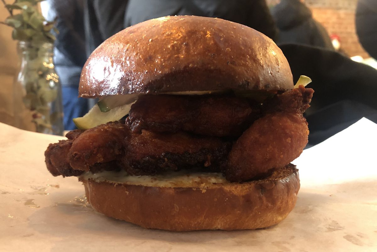 Dark pieces of fried chicken and green radish slices placed between two fluffy buns, sitting on a piece of white wax paper.