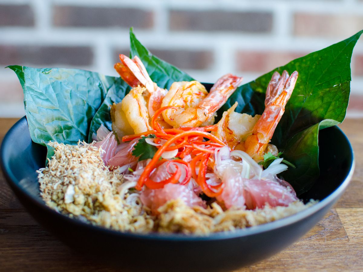 A Thai pomelo salad with shrimp, betel leaves, thinly sliced red chile, and a variety of crispy condiments sits in a black bowl on a wooden table in front of a brick background