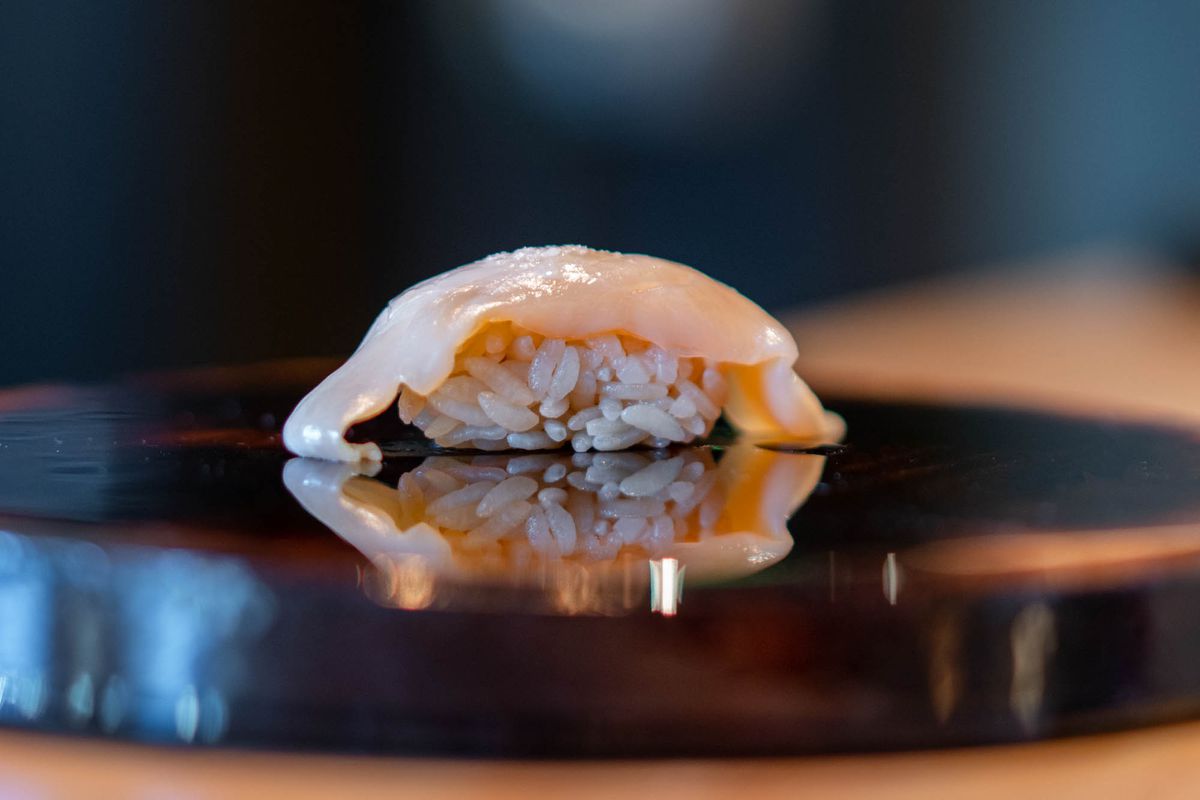 A piece of white-pink clam meat on rice on a black lacquered surface.