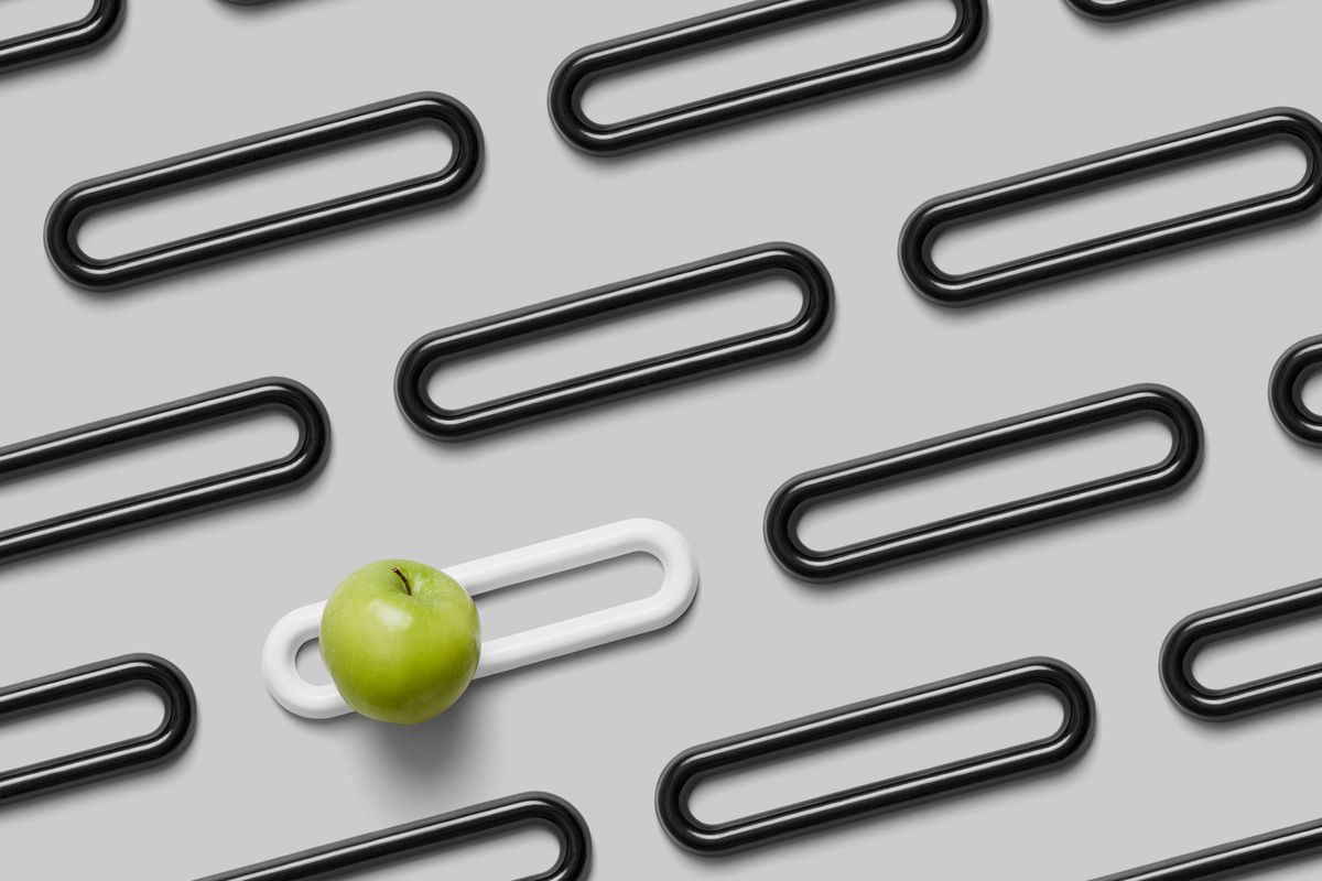 A series of simple elongated loops made of 3D-printed black porcelain arranged in a grid, with a white one with a single apple on top of it. 