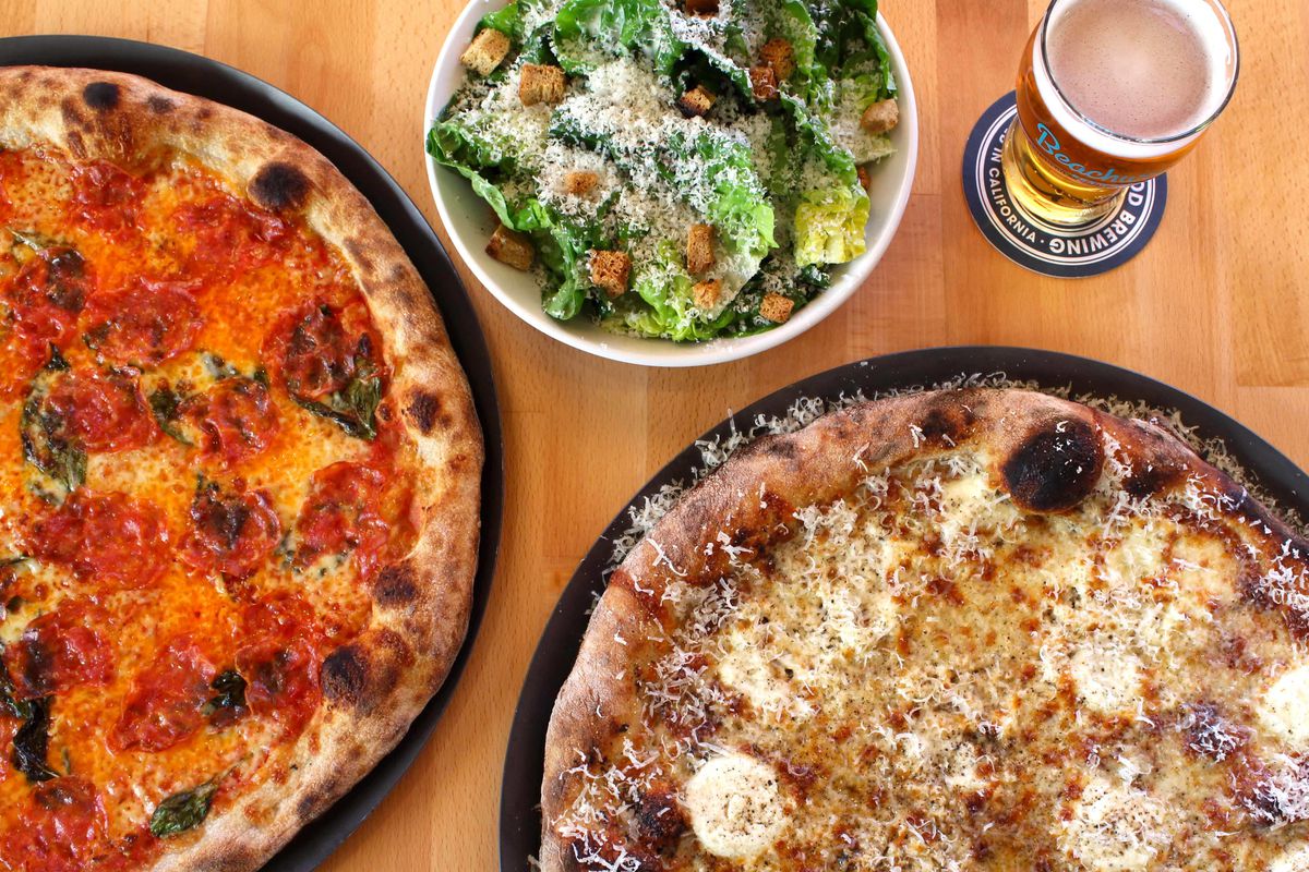 An overhead shot of a wooden table with two pizzas, a pint of golden ale, and a green salad.