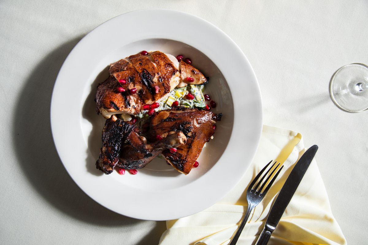 Pomegranate-lacquered grilled chicken with soft leeks and labneh on a white plate with fork and knife next to a wine glass.