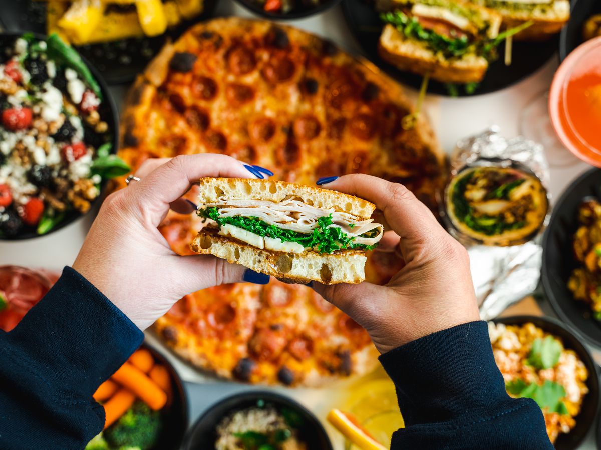 A first-person perspective of hands holding a sandwich in front of other food items like pizza, salads, burritos, and drinks. 