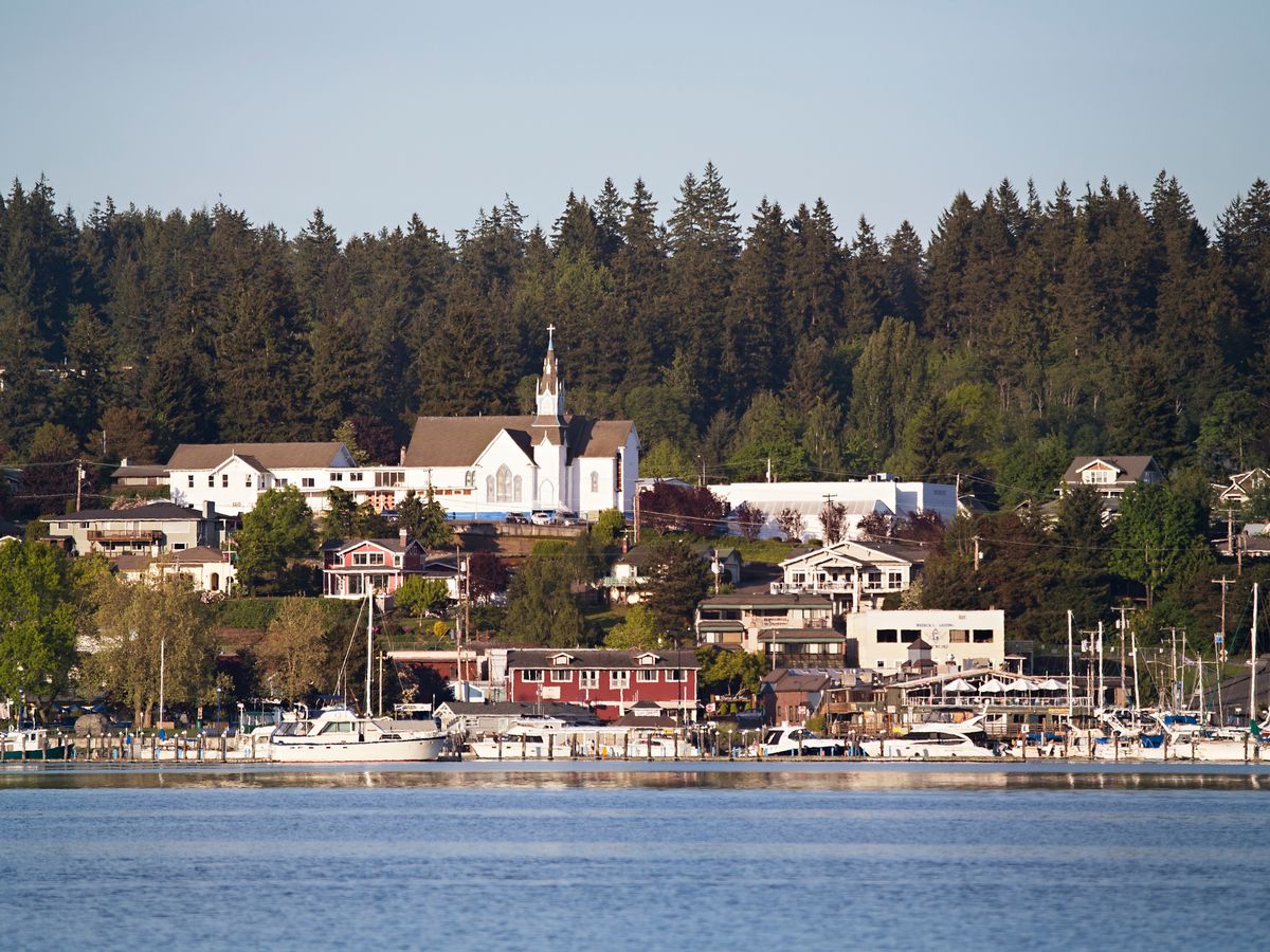 A seaside town, viewed from the water, includes a marina, small storefronts, and, up a hill past some trees, a white church with a steeple.
