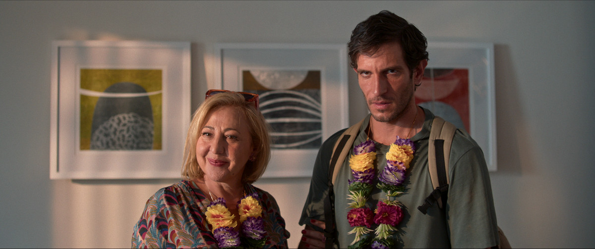 Husband (Carmen Machi) and adult son José Luís (Quim Gutiérrez) wearing flower lei necklaces in Honeymoon With My Mother.