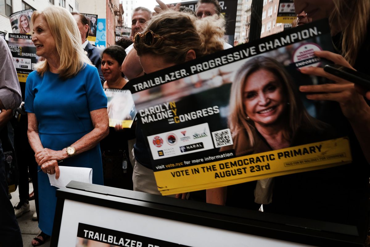 Maloney, blond haired in a blue dress, smiles amid supporters holding signs bearing her face that read “Trailblazer. Progressive Champion.”