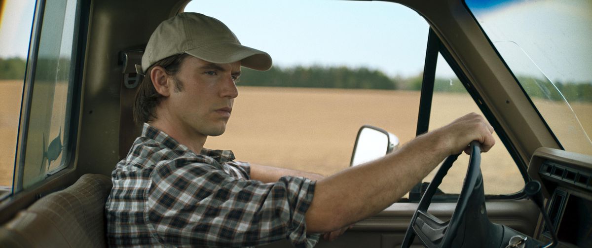 A young man in a checkered flannel shirt and a baseball cap sits behind the wheel of a flatbed truck while driving past a flat plain of dirt.
