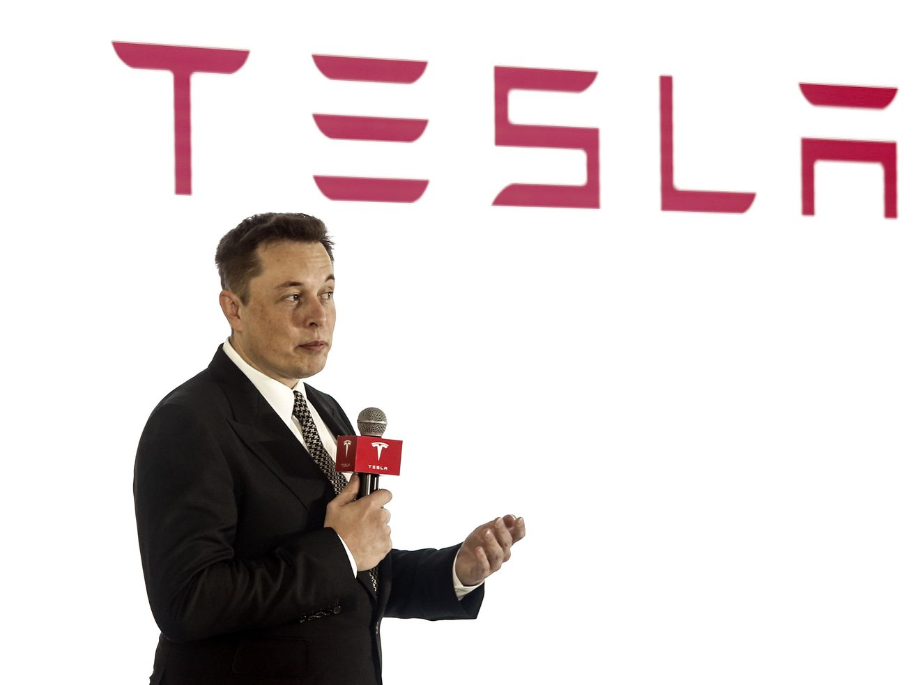 Elon Musk speaks into a handheld microphone, wearing a dark suit and tie and standing in front of a white backdrop with a large red Tesla logo. 