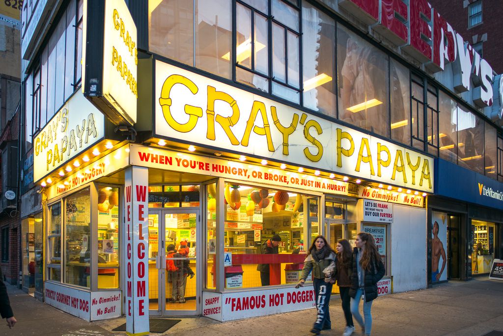 The Gray’s Papaya sign shines in its bright yellow color while patrons inside order hot dogs.