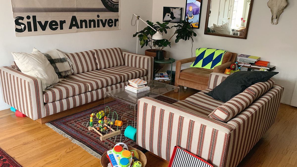 Two sofas in a striped red, green, and tan pattern face each other, with a coffee table in the middle, in a large room. A large poster hangs on the wall along with a mirror, two picture frames, and a steer skull. Baby toys are on the floor, near the rug.