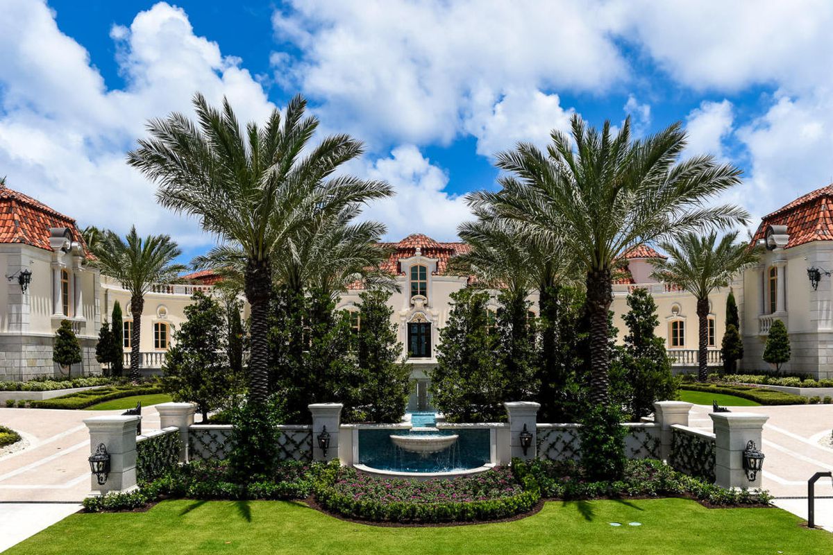 Front street view of a huge mansion in Palm Beach with palms, a dynamic driveway and a fountain out front.