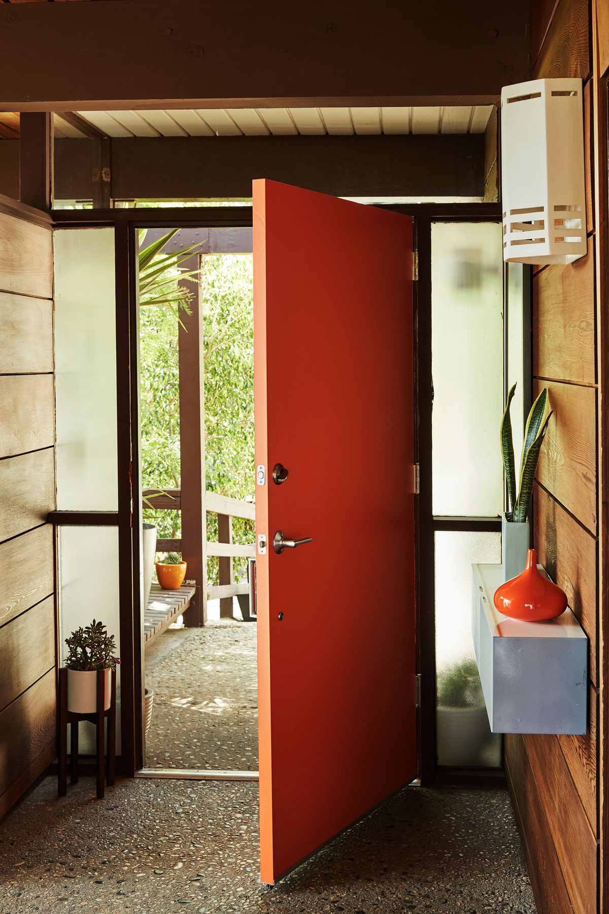 A front door painted red. The outside view is of a porch and trees. Inside, there is a shelf with a houseplant and a light fixture. 