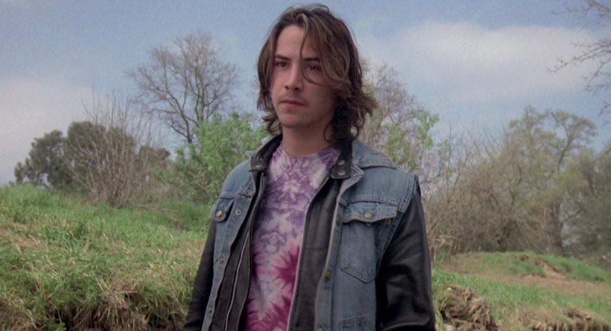 A young Keanu Reeves with long hair wears a denim jacket and a tie-die shirt against a background of grass and trees in River’s Edge.