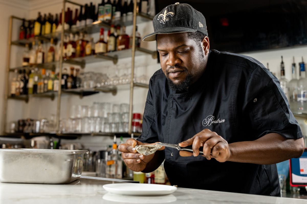 A man in a black chef’s jacket and black hat with a fleur de lis on it shucks oysters on a marble coutertop. Behind him are bar shelves with liquor.
