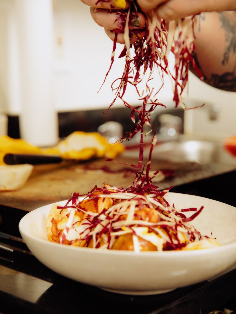 A plate of food is obscured by a handful of chopped radicchio being dropped from above as garnish.