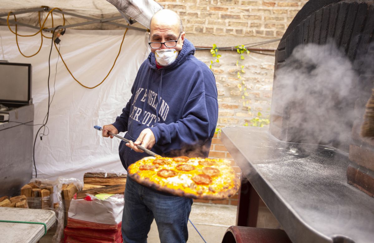 A man in a medical mask lifts a pizza out of an outdoor oven.