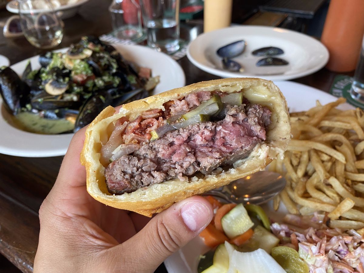 A medium rare burger is encased in fried dough. Mussels, french fries, and other dishes are visible in the background.