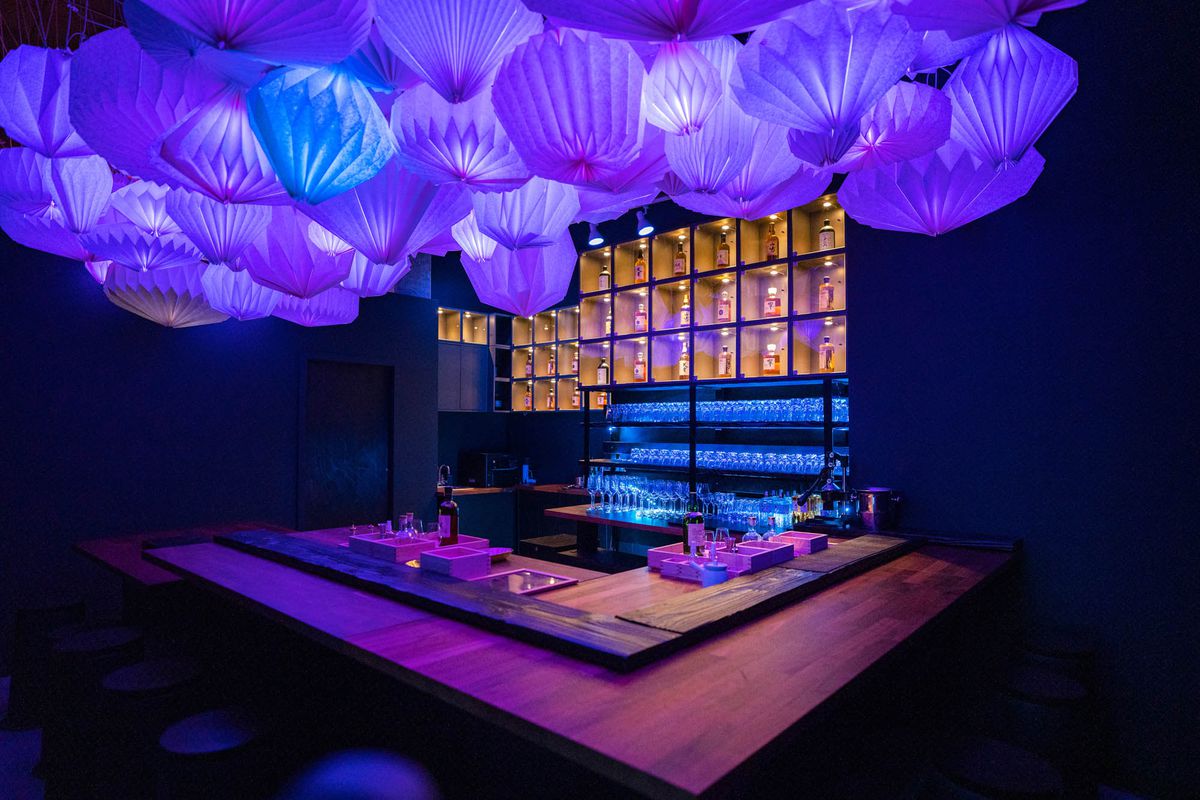 A square-shaped bar with lit-up bottles of whiskey behind it, and purple paper lanterns hanging above.